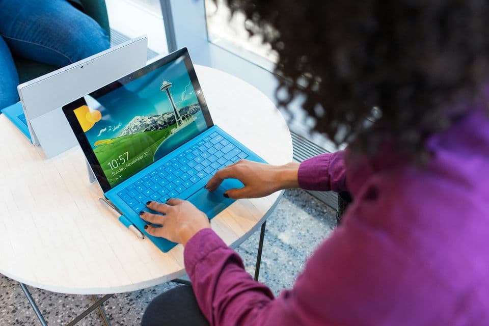 A person in a purple shirt typing on a bright blue laptop at a round table. The picture sits with this piece because a community bot is still someone at a keyboard, answering in a shared room.