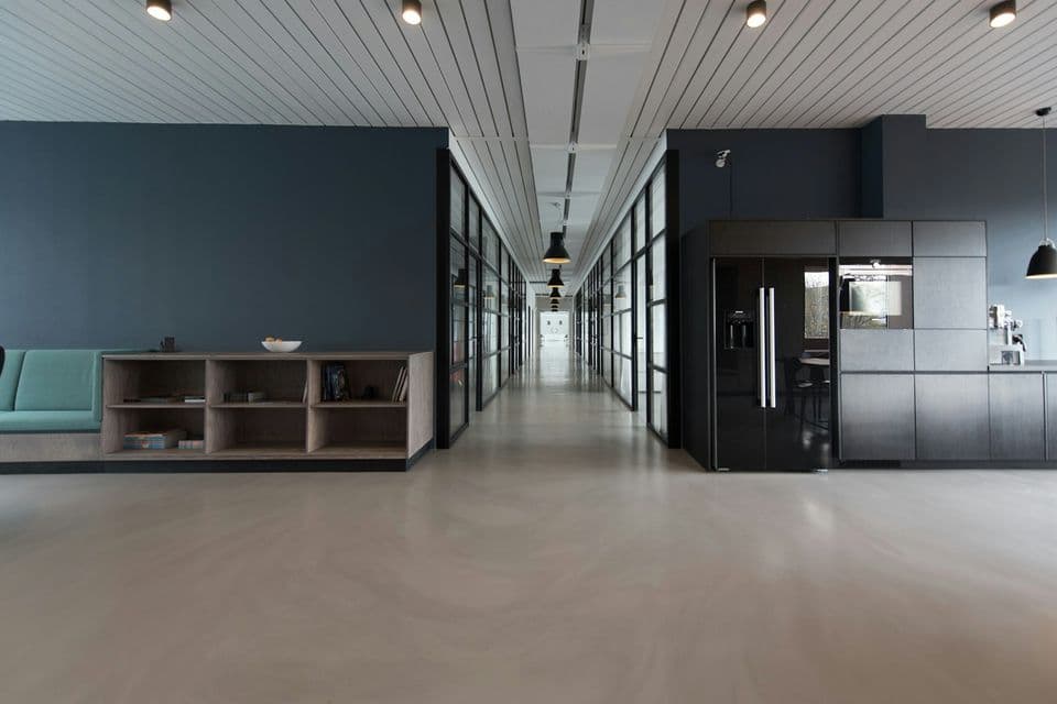 A long, quiet office corridor with glass rooms, a teal sofa, and a dark kitchen, a settled workplace for work that continues after a launch.