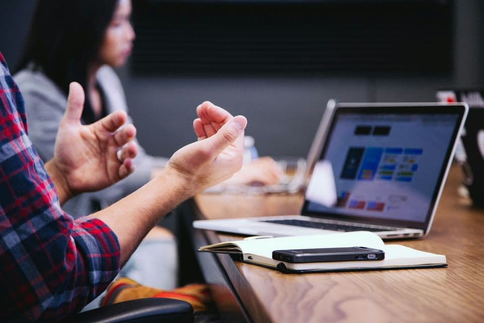 A person gesturing in a meeting, with a laptop showing a colorful layout, a notebook, and a phone on the table, a review where working habits become visible.