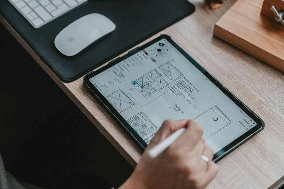 A hand holding a stylus over a tablet covered in wireframe sketches, with a keyboard and mouse beside it. The sketch is how a complex product becomes something a person can point at.