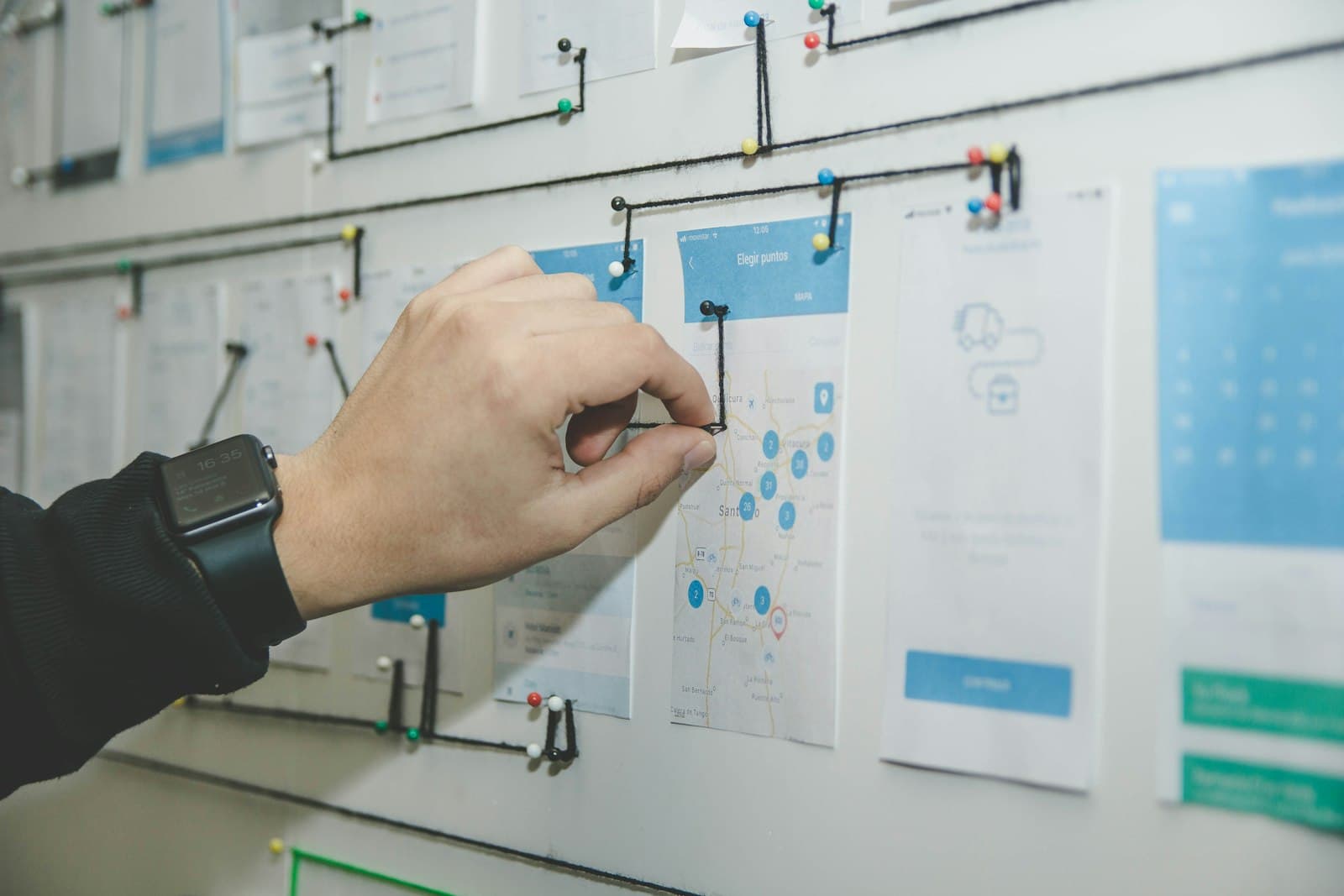 A hand pinning string between printed mobile screens on a wall, a physical map of how one screen leads to the next.