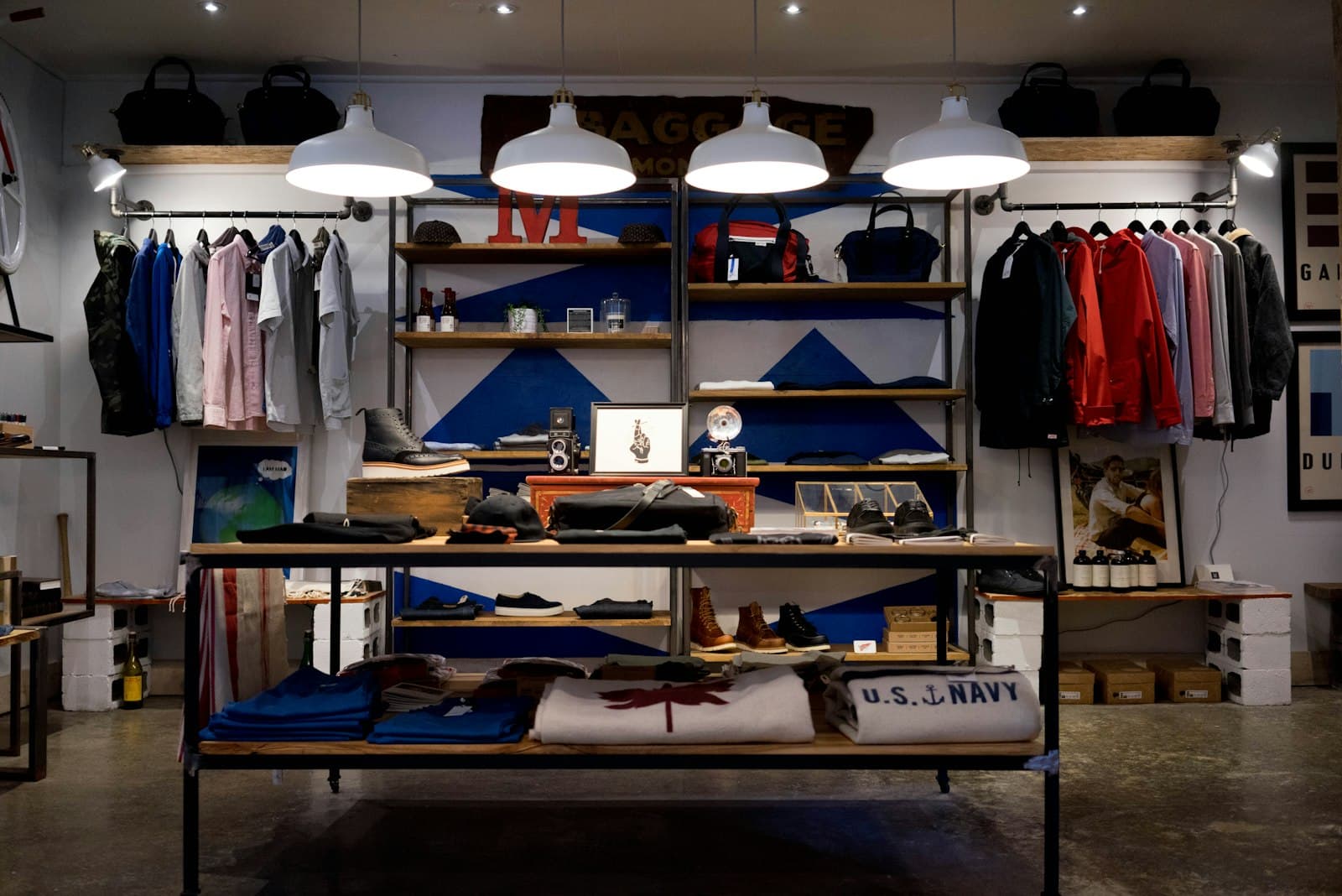A clothing shop interior with folded shirts, hanging jackets, shelves, and pendant lights. The room is edited: a few garments under a clear light, which is the shape of a brand shop.
