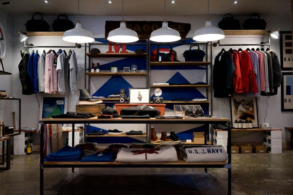 A clothing shop interior with folded shirts, hanging jackets, shelves, and pendant lights. The racks are the variant problem: many sizes and colors, one product the page still has to explain.
