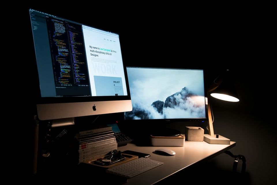 A dim desk with an iMac showing a code editor beside a portfolio page, a second monitor with a mountain wallpaper, and a lit lamp. The two screens fit a piece about the site a founder can open and the code behind it.