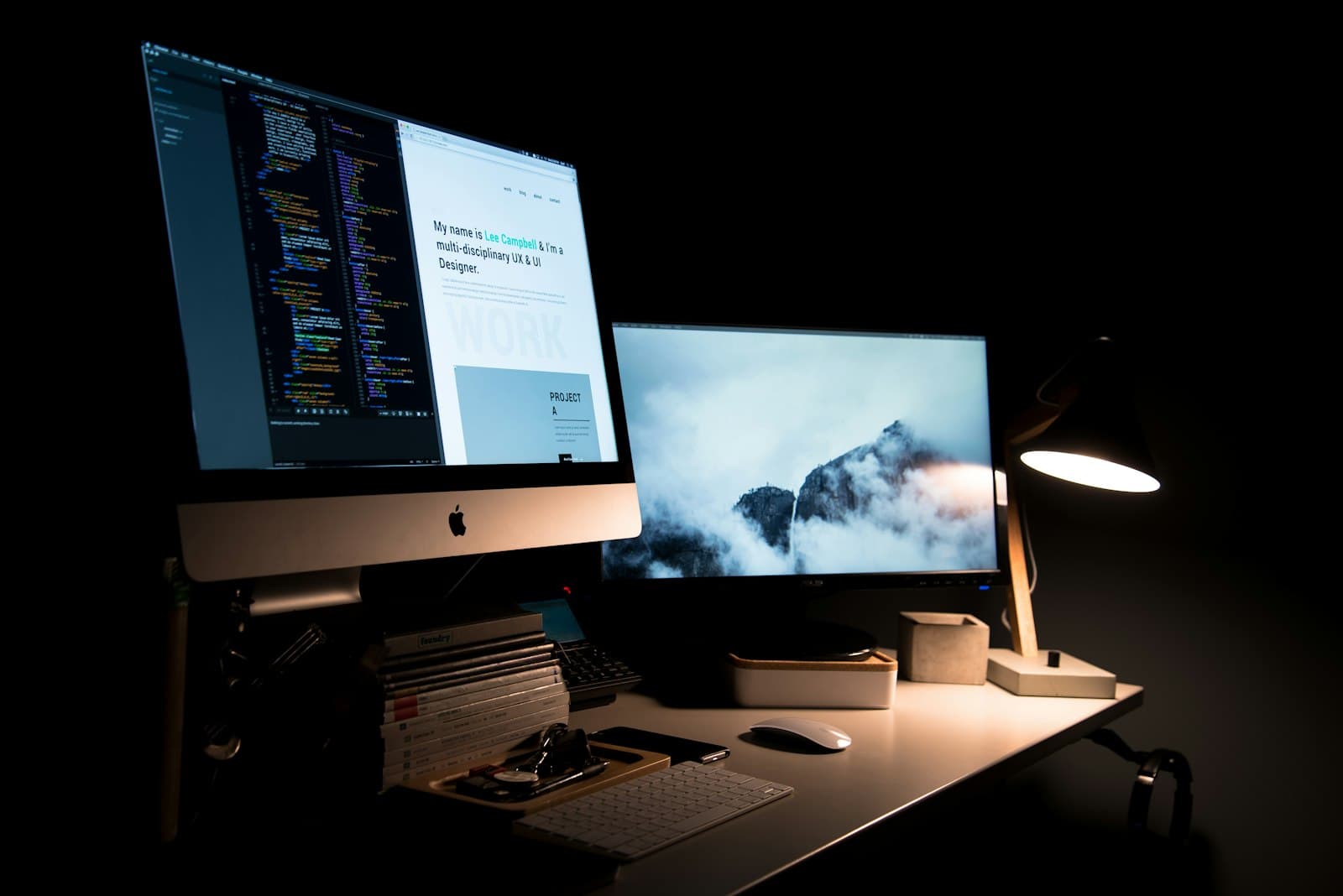 A dim desk with an iMac showing a code editor beside a portfolio page, a second monitor with a mountain wallpaper, and a lit lamp. The two screens fit a piece about the site a founder can open and the code behind it.