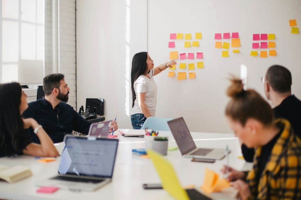 A workshop where one person points at colorful sticky notes while others sit with laptops. This is the working session you are hiring, not a pitch deck.